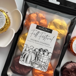 May include: A black bag filled with shower steamers wrapped in gold, copper, and brown foil. The label reads "Fall Festival Shower Steamers" with a graphic of pumpkins and a basket of fruit. The bag contains 8 oz (230g) of product.