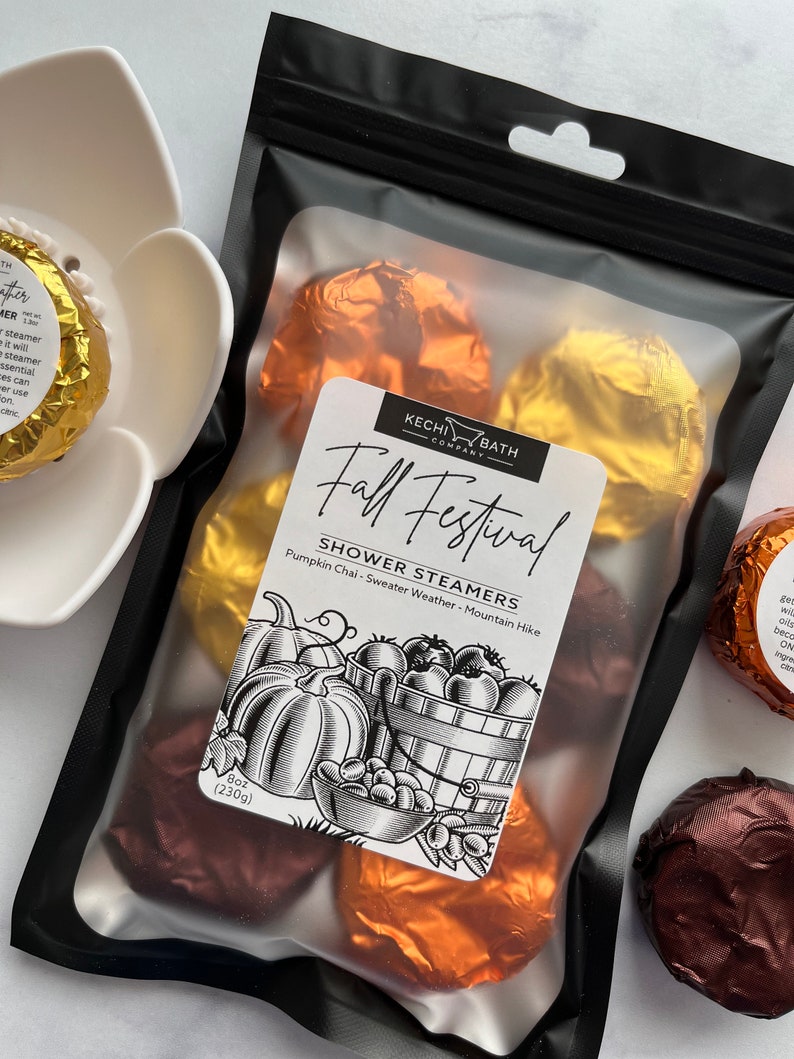 May include: A black bag filled with shower steamers wrapped in gold, copper, and brown foil. The label reads "Fall Festival Shower Steamers" with a graphic of pumpkins and a basket of fruit. The bag contains 8 oz (230g) of product.