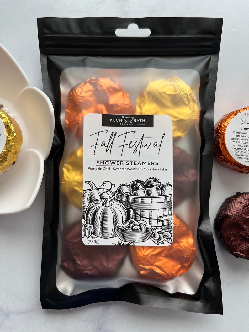 May include: A black package containing shower steamers. The label reads "Fall Festival Shower Steamers" with a pumpkin and apple illustration. The package contains several foil-wrapped steamers in shades of brown, gold, and orange. The package weighs 8 oz (230g).