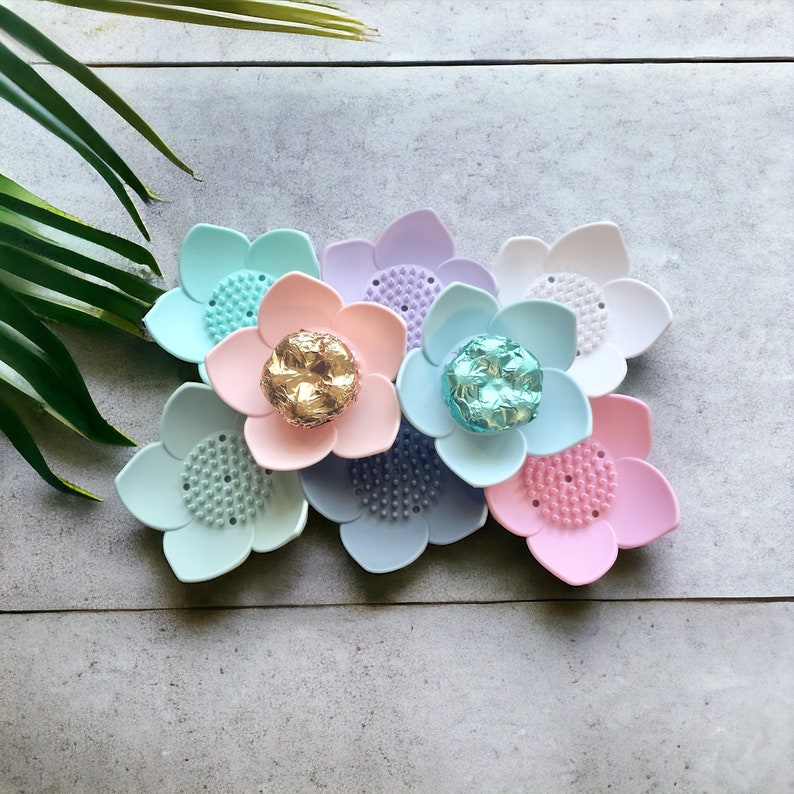 May include: A collection of colorful, flower-shaped soap dishes in pastel shades of blue, pink, purple, and white. Each dish has a textured center and a decorative, metallic accent in the center of some of the flowers.