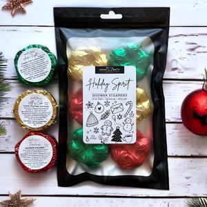 Puede incluir: Un paquete negro que contiene pastillas de ducha envueltas en papel de aluminio rojo, verde y dorado. El paquete tiene una etiqueta que dice "Holiday Spirit" con ilustraciones festivas. Contiene 230g de pastillas de ducha.
