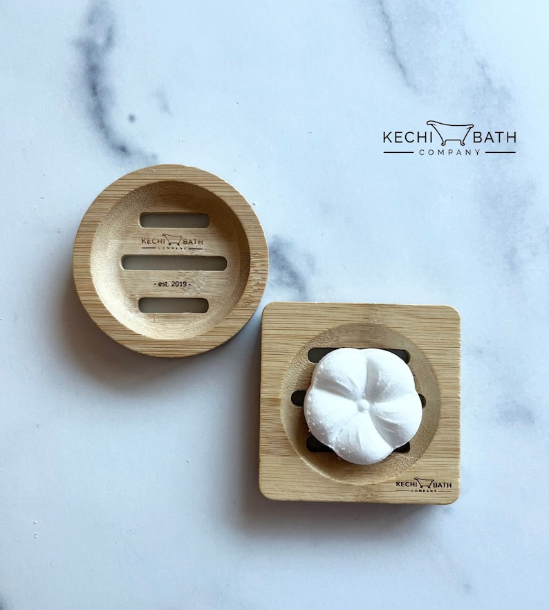 May include: Two bamboo soap dishes, one round and one square, with a white soap flower in the square dish. The round dish has the text "KECHI BATH COMPANY - est. 2019 -" printed on it.