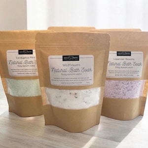 May include: Three brown paper bags filled with bath salts. The bags are labeled with the names of the scents: Eucalyptus Mint, Wildflowers, and Lavender Blooms. Each bag has a white label with the text "Natural Bath Soak" and "fizzy epsom salts".