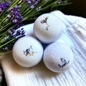 May include: Three white bath bombs with lavender and blue accents. The bath bombs are on a white towel.