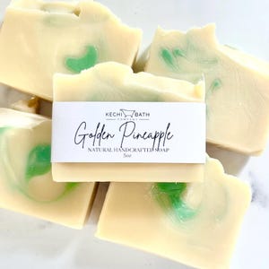 May include: Several bars of natural handcrafted soap with a creamy yellow base and green swirls. A white label on one bar reads "Golden Pineapple" and "5oz". The soap is from Kechi Bath Company.