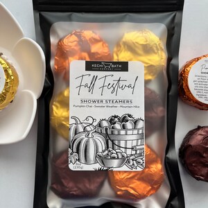 May include: A black package containing shower steamers. The label reads "Fall Festival" with a pumpkin and apple illustration. The package contains several foil-wrapped shower steamers in shades of brown, gold, and orange. The package weighs 8 oz (230g).
