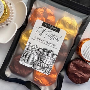 May include: A clear plastic bag containing several shower steamers wrapped in gold and copper foil. The bag has a label that reads "Fall Festival" and lists the scents: Pumpkin Chai, Sweater Weather, and Mountain Hike. The bag contains 8 oz (230g) of product.