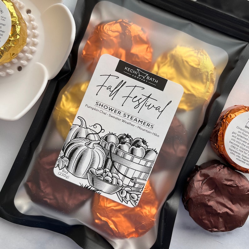 May include: A clear plastic bag containing several shower steamers wrapped in gold and copper foil. The bag has a label that reads "Fall Festival" and lists the scents: Pumpkin Chai, Sweater Weather, and Mountain Hike. The bag contains 8 oz (230g) of product.