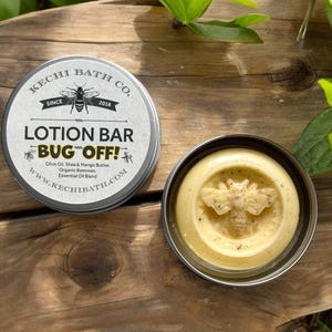 May include: A round tin container with a white label that reads "KECHI BATH CO. SINCE 2018 LOTION BAR BUG OFF! Olive Oil, Shea & Mango Butter. Organic Beeswax. Essential Oil Blend WWW.KECHIBATH.COM". The container is open and shows a yellow lotion bar with a bee design.
