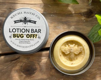 May include: A round tin container with a white label that reads "KECHI BATH CO. SINCE 2018 LOTION BAR BUG OFF! Olive Oil, Shea & Mango Butter. Organic Beeswax. Essential Oil Blend WWW.KECHIBATH.COM". The container is open and shows a yellow lotion bar with a bee design.