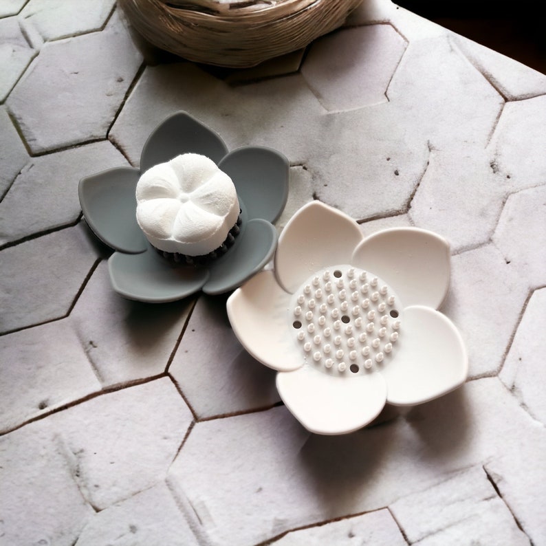 May include: Two gray and white flower-shaped soap dishes. One dish has a white soap bar in the center. The other dish has a textured surface.
