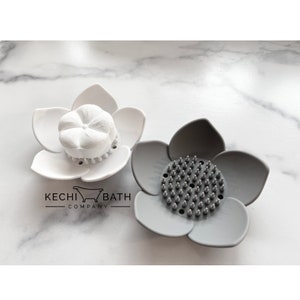 May include: Two flower-shaped soap dishes, one white and one gray. The white dish has a round soap bar with a flower design in the center. The gray dish has a textured surface with small bumps.
