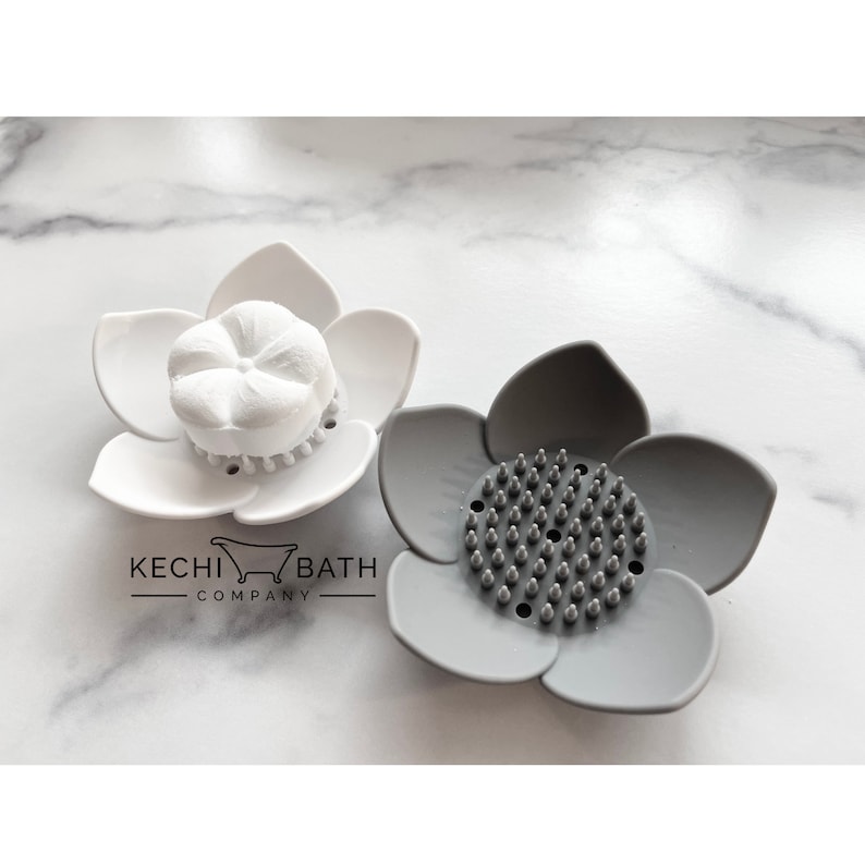 May include: Two flower-shaped soap dishes, one white and one gray. The white dish has a round soap bar with a flower design in the center. The gray dish has a textured surface with small bumps.