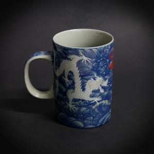May include: A ceramic mug with a white interior and handle. The exterior features a blue and white wave pattern with a white dragon design. A red accent is visible on the side of the mug.