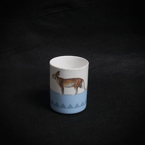 May include: A white ceramic mug featuring a leopard illustration. The mug has a light blue base with a geometric pattern and a distressed rim. The leopard is brown with black spots.