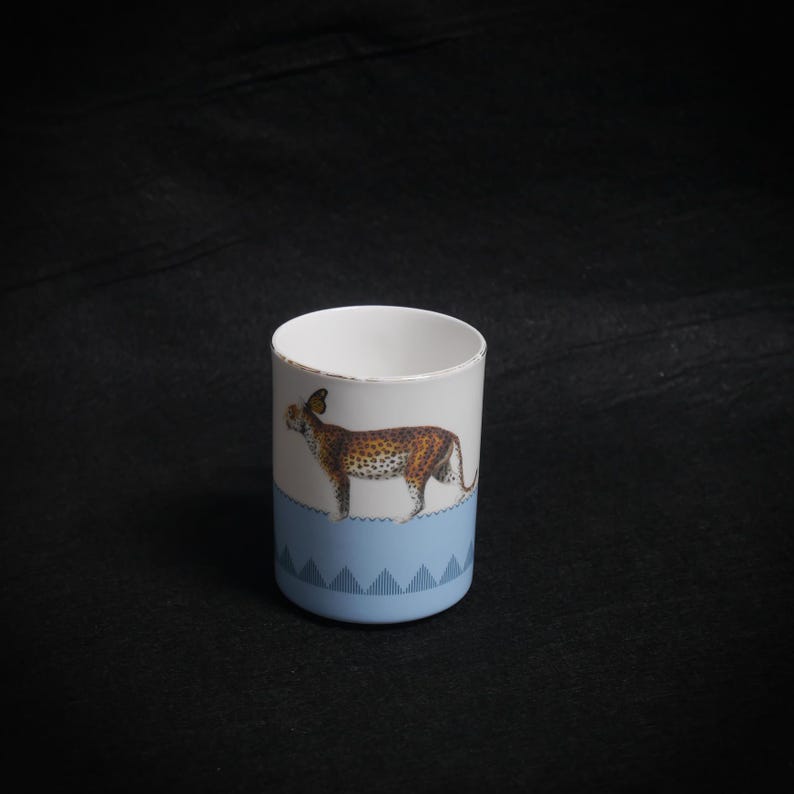 May include: A white ceramic mug featuring a leopard illustration. The mug has a light blue base with a geometric pattern and a distressed rim. The leopard is brown with black spots.