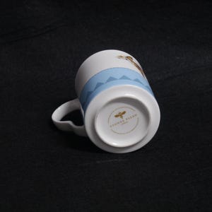 May include: White ceramic mug with a light blue band featuring a stitched design. The mug has a curved handle and a printed design of a giraffe. The bottom of the mug has the text "YVONNE ELLEN LONDON".