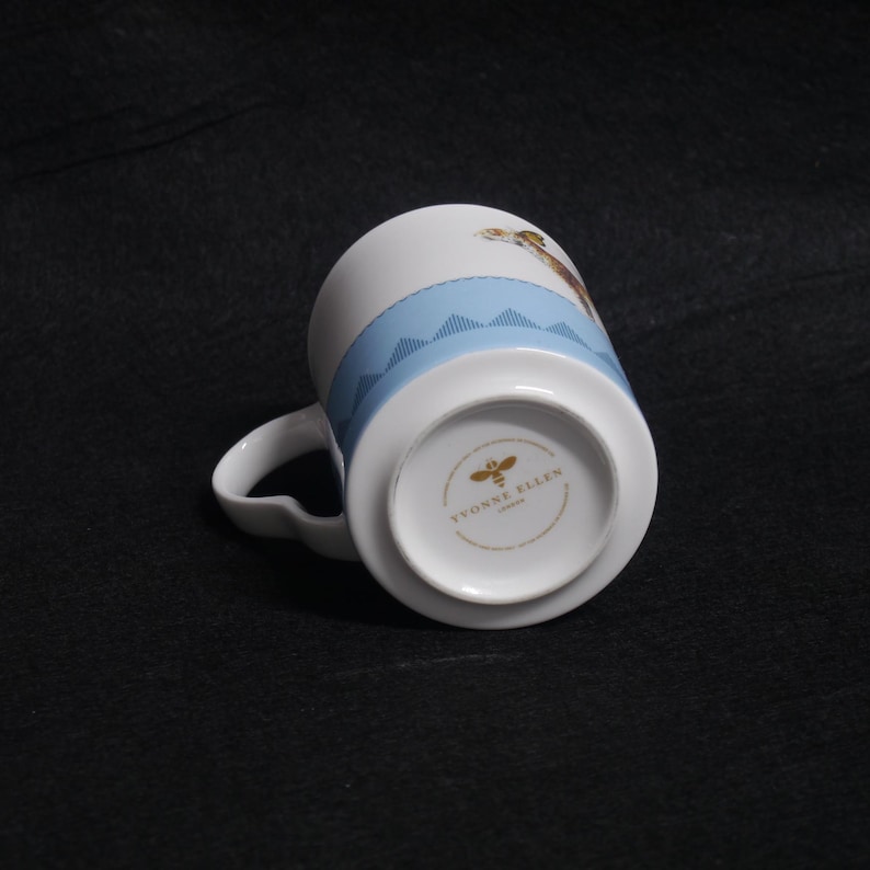 May include: White ceramic mug with a light blue band featuring a stitched design. The mug has a curved handle and a printed design of a giraffe. The bottom of the mug has the text "YVONNE ELLEN LONDON".