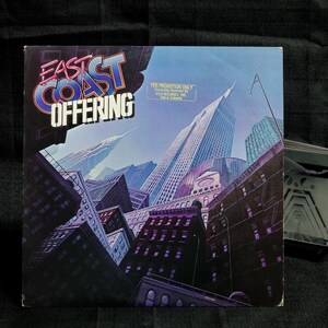 May include: A vintage vinyl record cover for the band East Coast Offering. The cover features a cityscape with tall buildings and a blue sky. The band's name is printed in large, bold letters in the upper left corner. The text "For Promotion Only" is printed in the upper right corner.