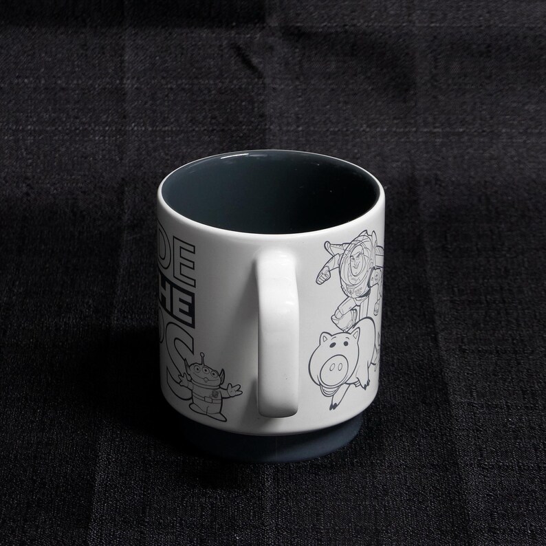 May include: White ceramic mug with a black interior and a black and white outline drawing of characters from the Toy Story movie franchise. The text "To infinity and beyond" is printed on the mug.