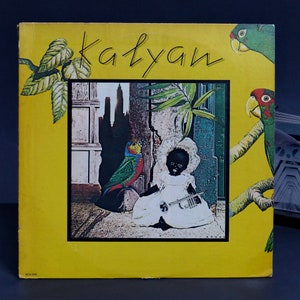 May include: A yellow vinyl record cover with the title "Katyan" in black cursive lettering. The cover features a collage of images including a child in a white dress holding a trumpet, a parrot, and a cityscape.