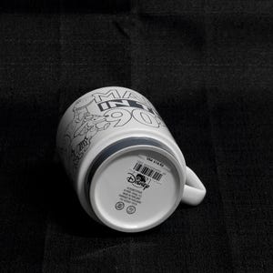 May include: White ceramic mug with a black and white illustration of characters from the Disney movie "Monsters Inc." The mug has the text "Monsters Inc. 90s" on the front. The bottom of the mug has a gray band and the Disney logo.