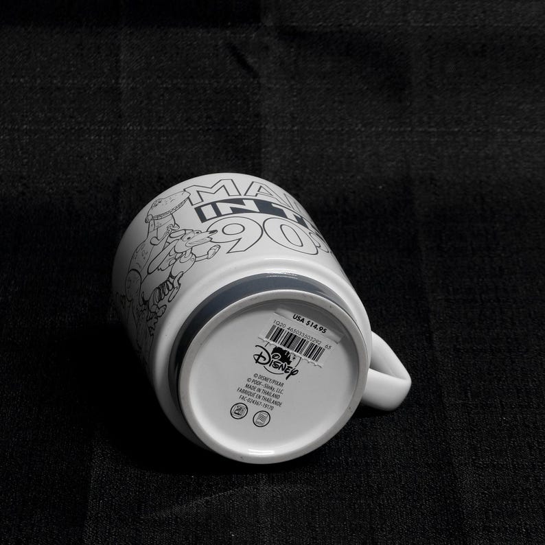 May include: White ceramic mug with a black and white illustration of characters from the Disney movie "Monsters Inc." The mug has the text "Monsters Inc. 90s" on the front. The bottom of the mug has a gray band and the Disney logo.