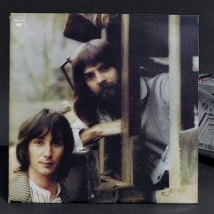 May include: A vintage vinyl record album cover featuring two men with long hair. The men are standing in front of a wooden structure. The album cover is in a muted color palette of brown, beige, and white.