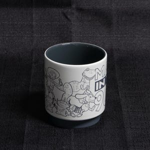 May include: White ceramic mug with a black and white illustration of characters from the Toy Story franchise. The mug has a black rim and a black interior. The text "Made in 90s" is printed on the side of the mug.