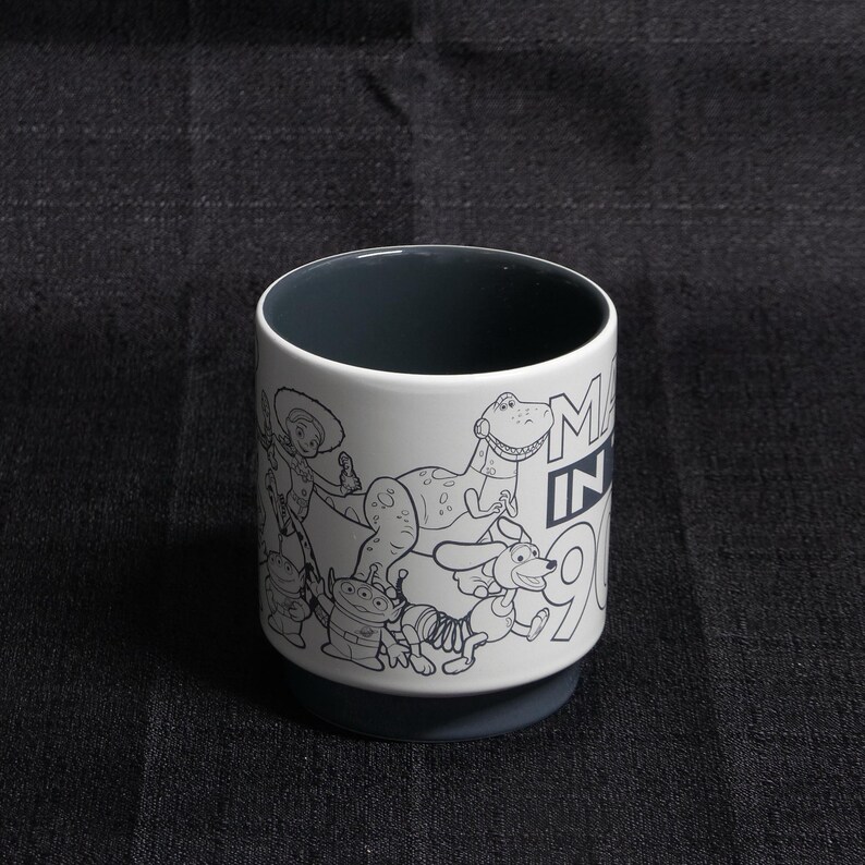 May include: White ceramic mug with a black and white illustration of characters from the Toy Story franchise. The mug has a black rim and a black interior. The text "Made in 90s" is printed on the side of the mug.