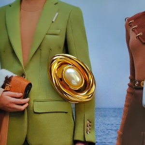 Può includere: Un broche ovale dorato con una grande perla finta al centro. Il broche ha un design testurizzato e a spirale.