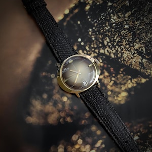 Op de afbeelding: Een gouden polshorloge met een zwarte leren band. De wijzerplaat van het horloge heeft een bruine gradiënt en de woorden "Sportus" en "Skymaster" staan erop gedrukt.