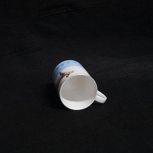 May include: White ceramic mug with a handle, featuring a design of a lizard and geometric blue pattern. The mug has a gold rim and is set against a dark background.