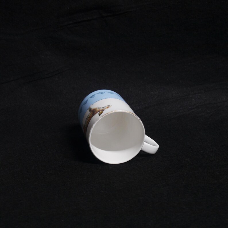 May include: White ceramic mug with a handle, featuring a design of a lizard and geometric blue pattern. The mug has a gold rim and is set against a dark background.