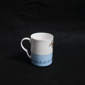 May include: White ceramic mug with a light blue band featuring a geometric pattern. The mug has a gold rim and a handle. A brown and white animal illustration is on the side of the mug.