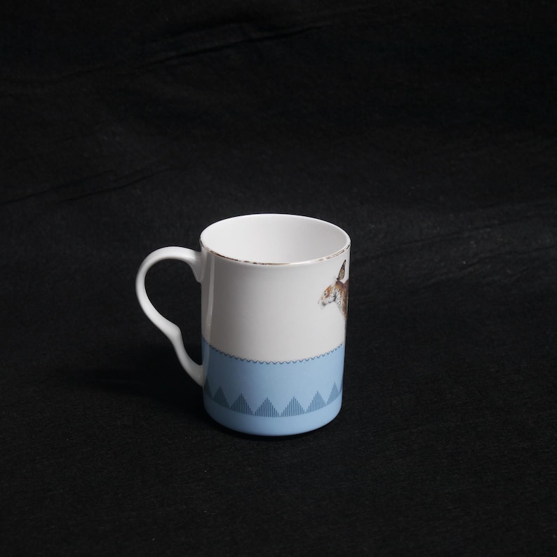 May include: White ceramic mug with a light blue band featuring a geometric pattern. The mug has a gold rim and a handle. A brown and white animal illustration is on the side of the mug.