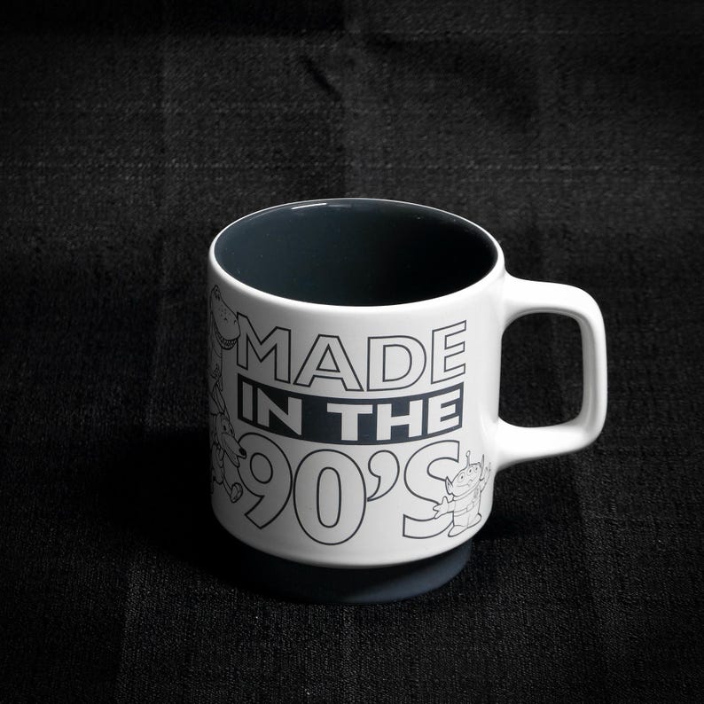 May include: White ceramic mug with a black interior and a black and white graphic that says "MADE IN THE 90'S". The graphic includes cartoon characters.