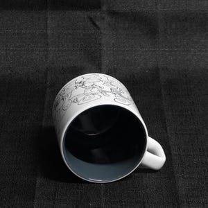 May include: A white ceramic mug with a black and white cartoon design. The mug has a black interior and a handle.