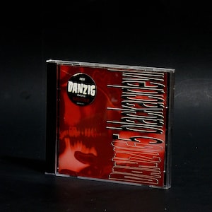 May include: A black CD case with a red and white design. The front of the case features the text "Danzig" and "Sacrifice" in white lettering. The back of the case features the text "Danzig" in white lettering.