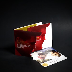 May include: A red and gold CD case with the text "Elvis Presley Christmas Duets" on the front. The case is open to reveal a CD with a picture of Elvis Presley on the front. The CD is titled "Where Elvis Lives" and has a picture of Elvis Presley on the front. The CD is in a clear plastic case. The CD case is on a black surface.