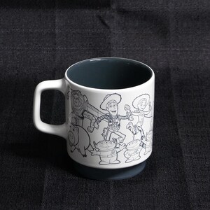 May include: White ceramic mug with a black interior and a black and white outline image of characters from the Toy Story franchise. The characters include Woody, Buzz Lightyear, and other toys.
