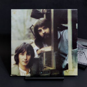 May include: A vintage vinyl record album cover featuring two men with long hair. The men are standing in front of a wooden structure. The album cover is in a brown and green colour scheme.