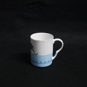 May include: White ceramic mug with a light blue base and a gold rim. The mug features a brown animal illustration and a decorative blue border with triangle patterns. The handle is white.