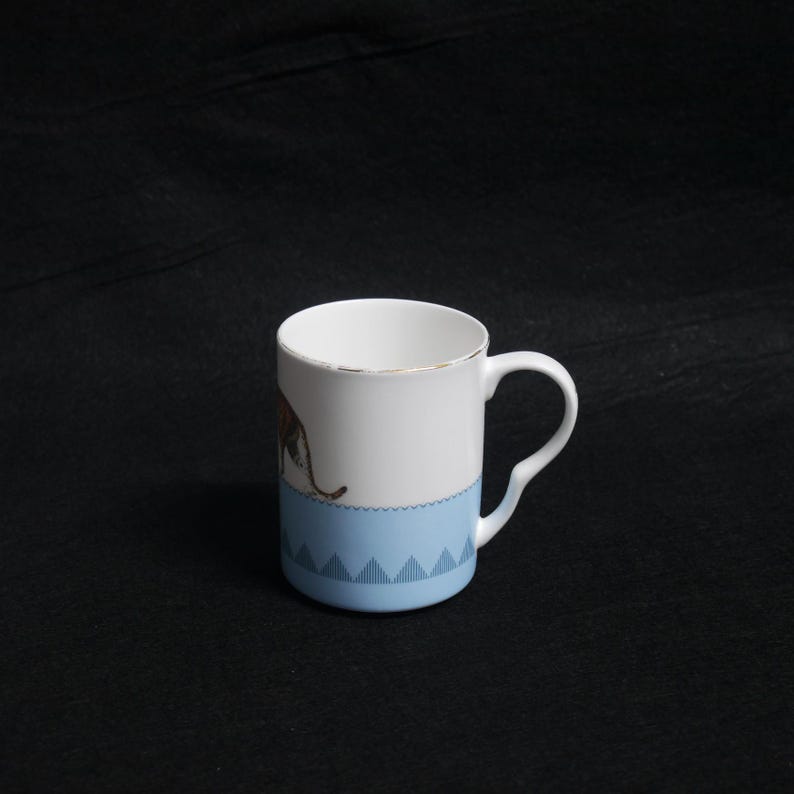 May include: White ceramic mug with a light blue base and a gold rim. The mug features a brown animal illustration and a decorative blue border with triangle patterns. The handle is white.