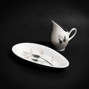 May include: A white oval serving dish with a black and gold floral design. A white creamer with a black and gold floral design sits next to the dish.