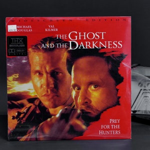 Puede incluir: Una caja de cinta VHS para la película "El fantasma y las tinieblas" protagonizada por Michael Douglas y Val Kilmer. La caja es roja con una imagen en blanco y negro de los actores en la parte delantera. El texto "Presa para los cazadores" está impreso en la parte inferior de la caja.