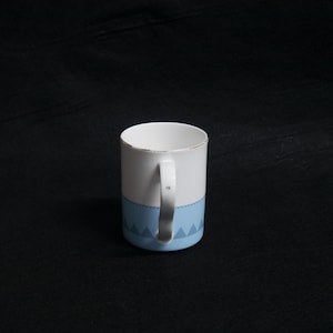 May include: White ceramic mug with a light blue geometric pattern at the base and a gold rim. The mug has a white handle and is set against a black background. The mug is designed for hot beverages.