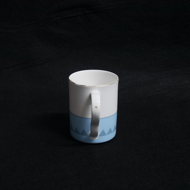 May include: White ceramic mug with a light blue geometric pattern at the base and a gold rim. The mug has a white handle and is set against a black background. The mug is designed for hot beverages.