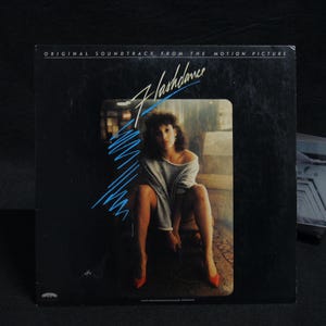 May include: A vinyl record album cover for the movie "Flashdance." The cover is black with the movie title in a stylized font and a photo of a woman in a gray sweater and red heels. The text "ORIGINAL SOUNDTRACK FROM THE MOTION PICTURE" is at the top.