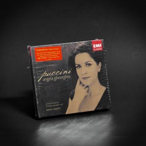 Puede incluir: Un CD de Puccini con Angela Gheorghiu, envuelto en plástico. La portada presenta un retrato de una mujer, con los textos "Puccini" y "Angela Gheorghiu". El CD está etiquetado como "EMI Classics" y "Gramophone 'Editors Choice'".